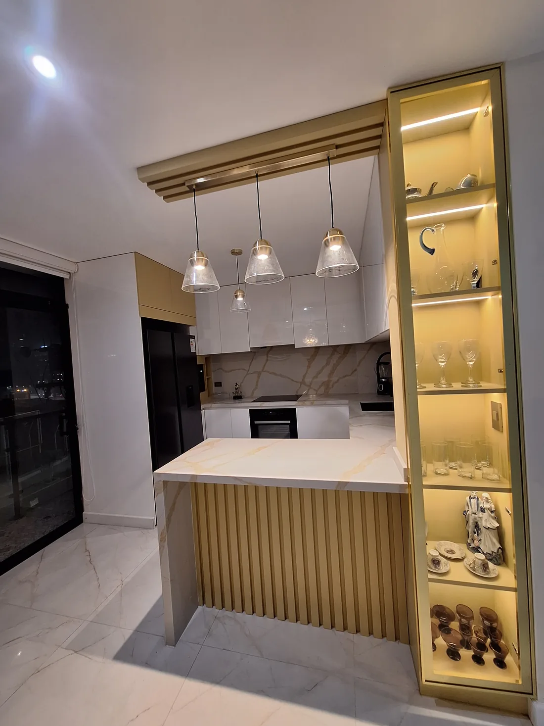 Cocina remodelada con muebles blancos, barra de mármol, listones dorados y vitrina iluminada