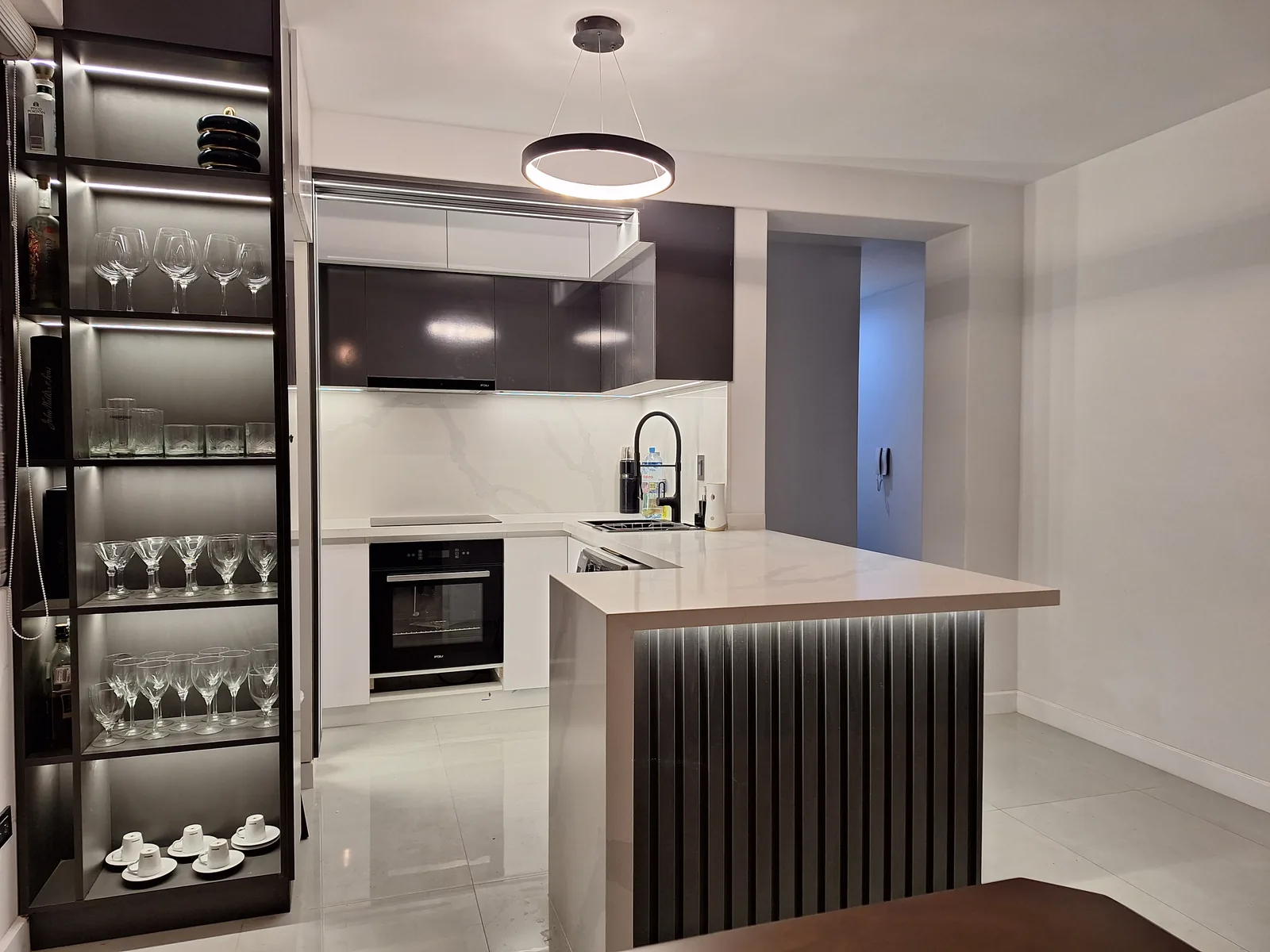 Cocina remodelada en negro y blanco con barra de listones iluminada y estantería de cristalería