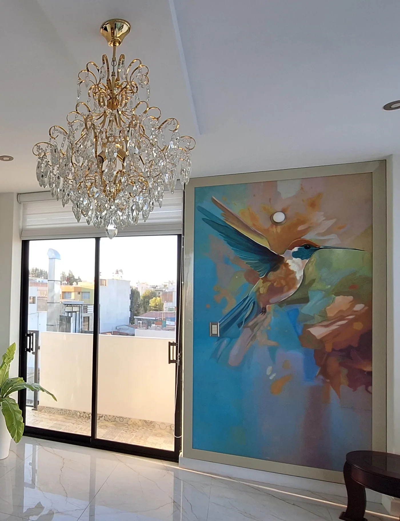 Sala remodelada con araña de cristal dorada, mural de colibrí y piso de porcelanato